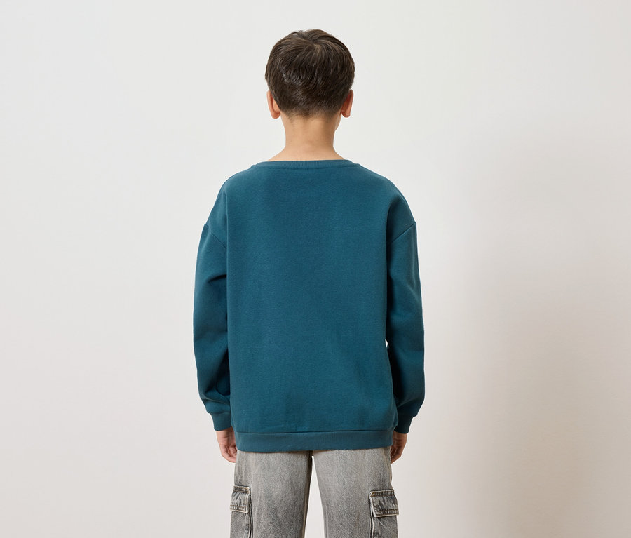 Pohľad zozadu na dieťa v modrozelenom svetri a sivých šortkách na bielom pozadí.