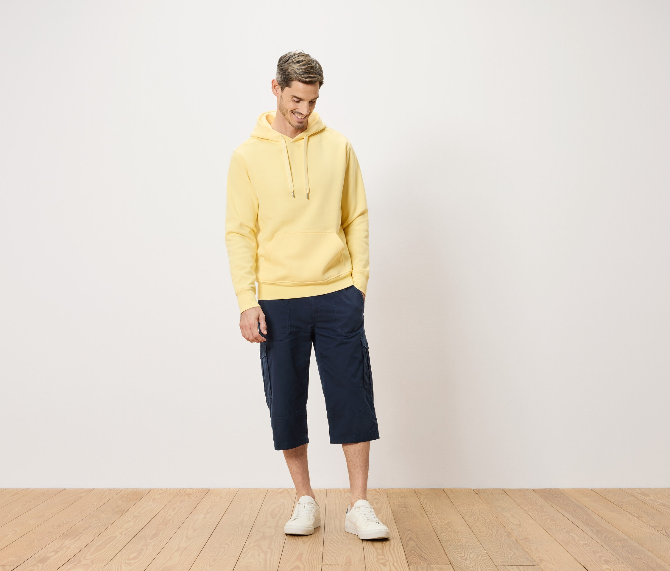 Muž v žltej mikine s kapucňou a modrých cargo šortkách stojaci na drevenej podlahe.