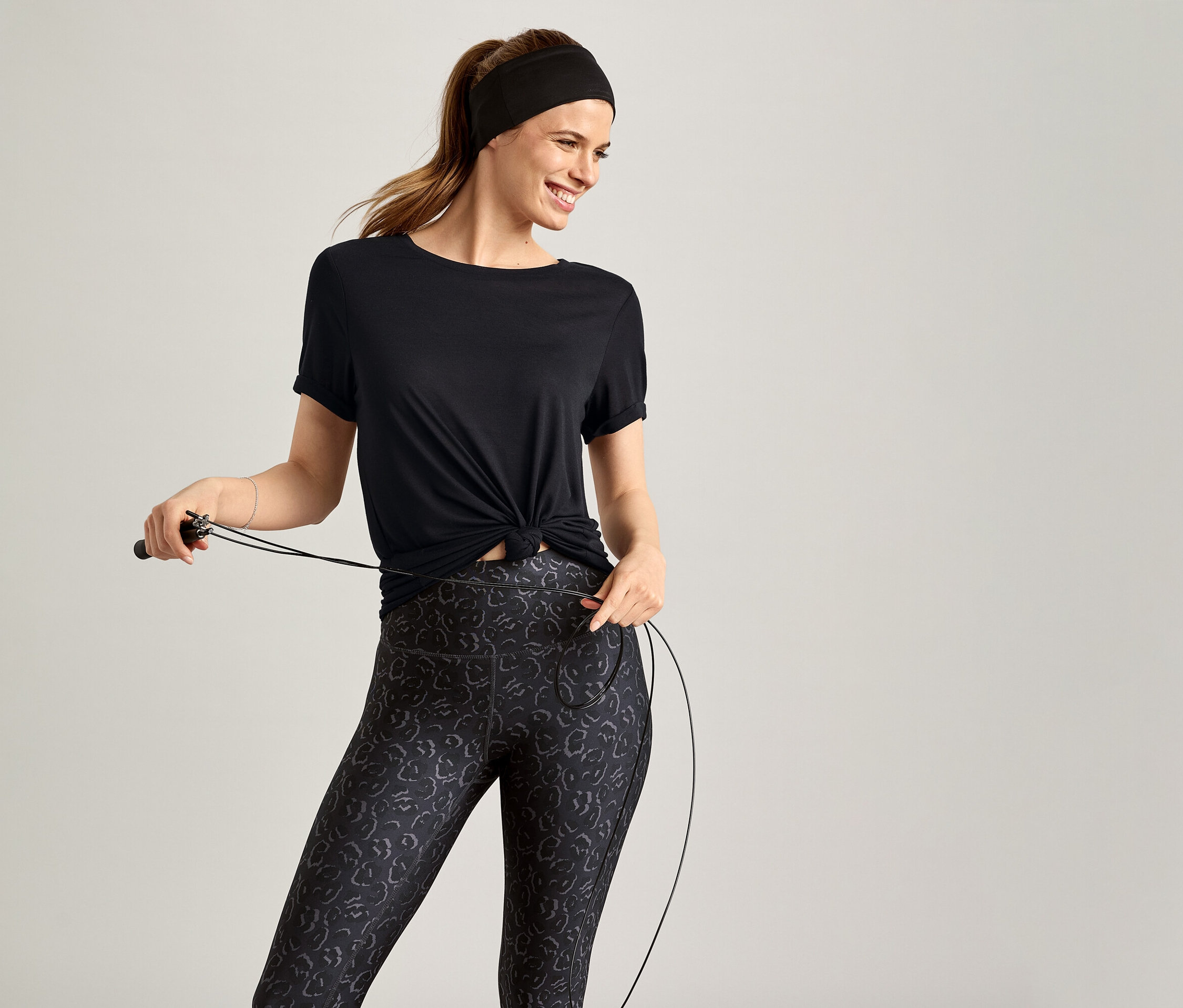Žena s čelenkou a v čiernom športovom tričku drží švihadlo na sivom pozadí.