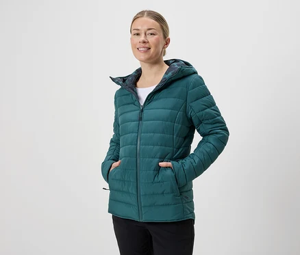Žena s kapucňou má na sebe smaragdovo zelenú prešívanú bundu na zips a čierne nohavice na neutrálnom pozadí.