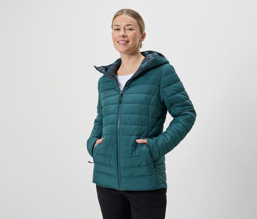 Žena s kapucňou má na sebe smaragdovo zelenú prešívanú bundu na zips a čierne nohavice na neutrálnom pozadí.