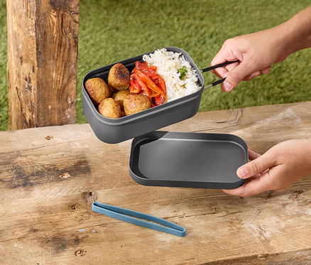 Ruky držia sivý obedár s ryžou, mäsovými guľkami a paprikou nad sivým vekom na drevenom stole.