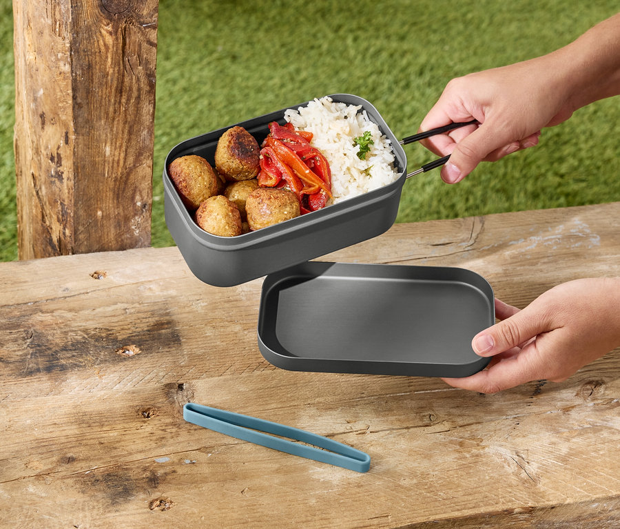 Ruky držia sivý obedár s ryžou, mäsovými guľkami a paprikou nad sivým vekom na drevenom stole.