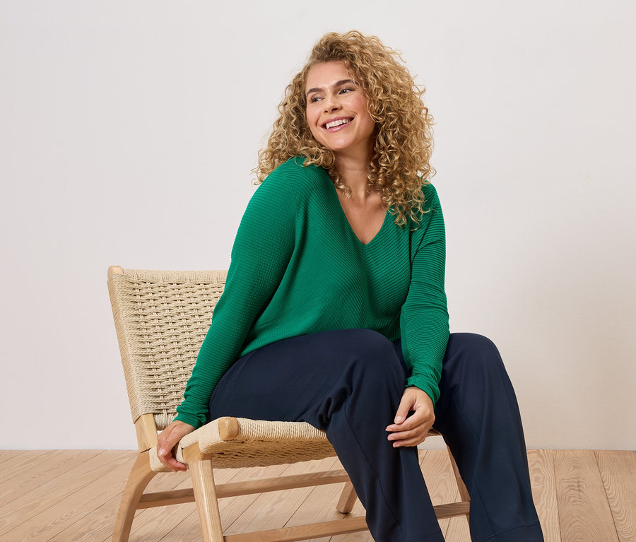 Žena s kučeravými vlasmi sedí na stoličke a má na sebe zelený sveter a modré nohavice.