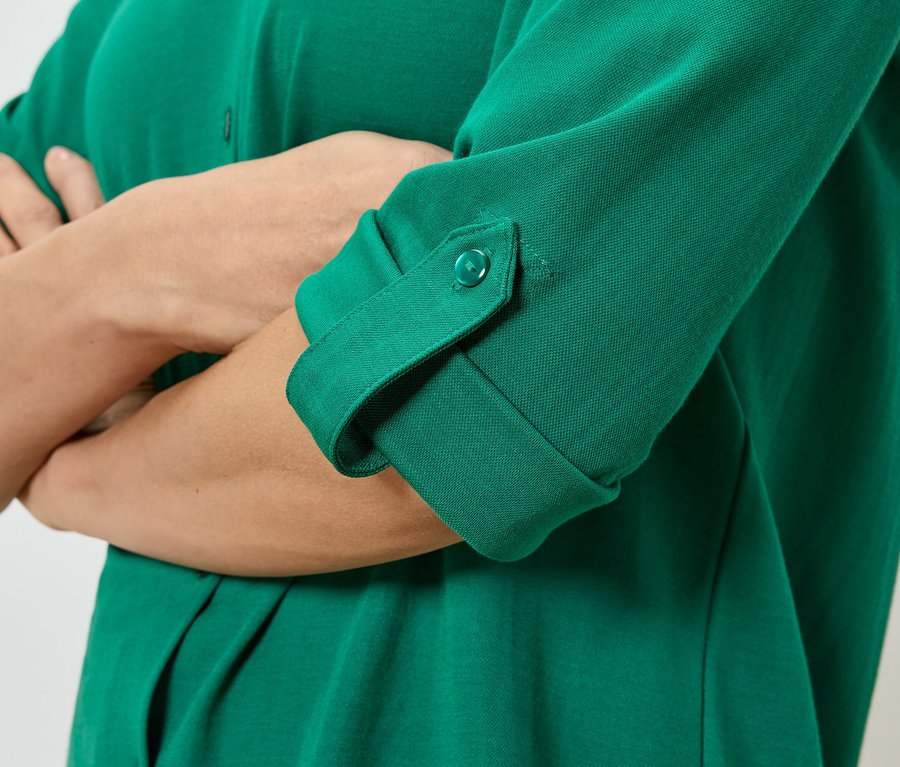 Detail osoby so skríženými rukami, ktorá má na sebe zelenú košeľu s gombíkmi a vyhrnutými rukávmi.
