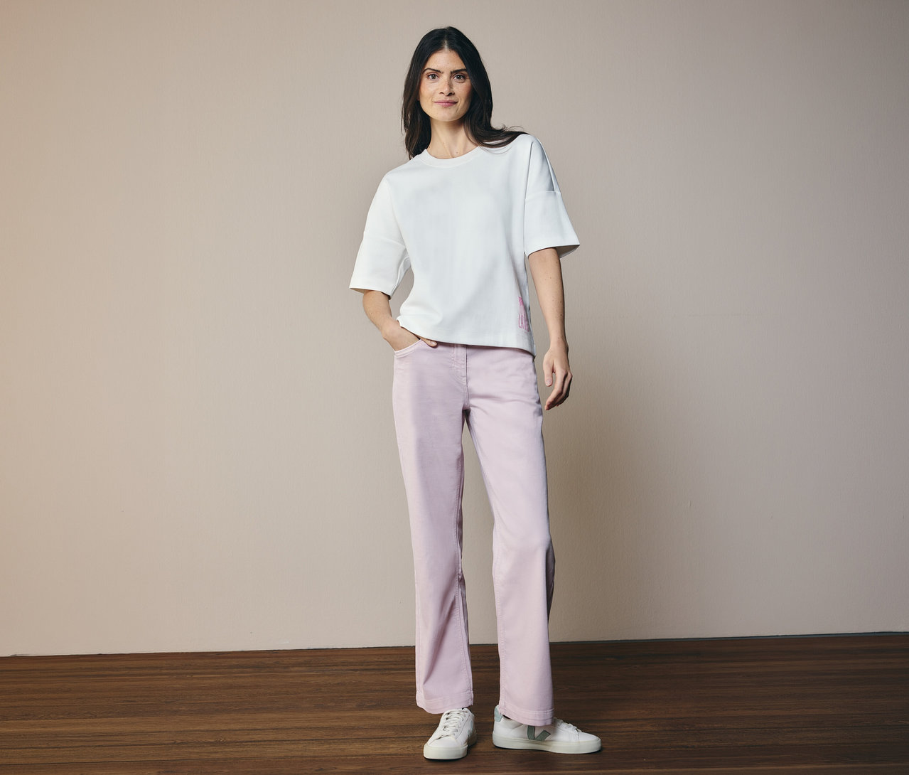 Žena v bielom tričku a džínsoch CECIL »Neele«. Stojí na drevenej podlahe pred béžovým pozadím.