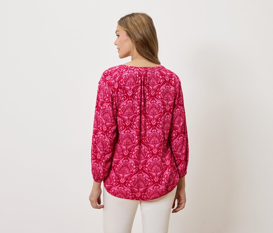 Pohľad zozadu na ženu s blond vlasmi, ktorá má na sebe ružovú blúzku s dlhým rukávom so vzorom a biele nohavice.