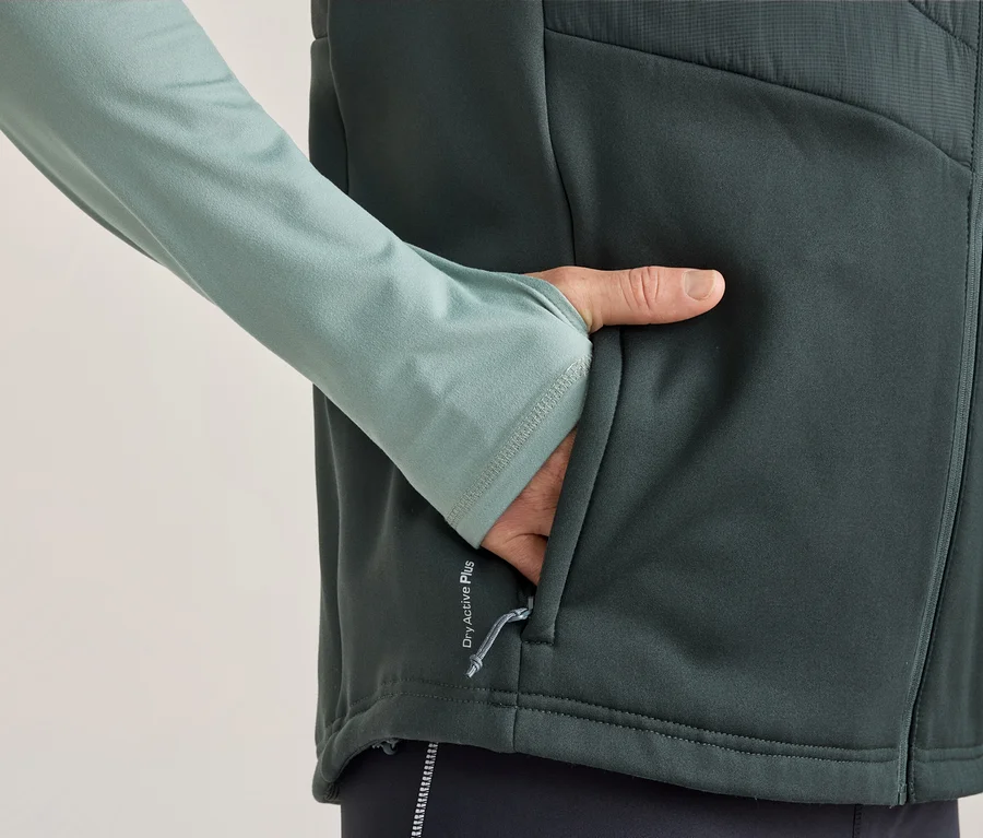 Osoba v svetlozelenom tričku s dlhým rukávom s otvormi na palce a tmavosivej veste, s rukou vo vrecku vesty. Na veste je nápis „Dry Active Plus“.