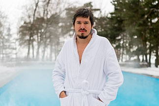 Bradatý muž v bielom župane stojí vonku pred parným bazénom s rukami vo vreckách.
