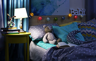 Mladá matka leží so svojím malým dieťaťom v posteli a číta mu z knihy, zatiaľ čo na nočnom stolíku svieti lampa.