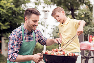 Mladý muž v zelenej zástere kľačí vedľa čierneho okrúhleho grilu, zatiaľ čo jeho syn kliešťami obracia mäsové špízy.