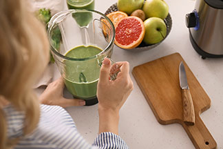 Kuchynské spotrebiče v akcii: žena stojí v kuchyni a v ruke drží svoj mixér, v ktorom je zelené smoothie – vedľa nej je miska ovocia a drevená doska na krájanie s nožom.