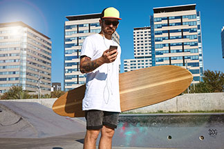 Skejter so slnečnými okuliarmi a šiltovkou stojí v tričku v nadrozmernom štýle so svojím longboardom v ruke v skejtparku a pozerá sa do svojho mobilu.