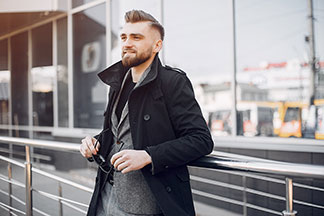 Pánska bunda do kancelárie: mladý muž v krátkom čiernom kabáte so sivým sakom pod ním sa opiera o zábradlie a drží v ruke slnečné okuliare.