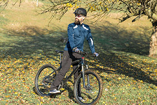 Bradatý muž na horskom bicykli vo funkčných teplákoch, tričku, prilbe a slnečných okuliaroch stojí za slnečného dňa na svahu a vychutnáva si výlet.