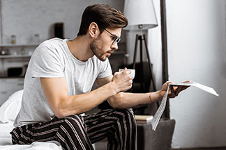Mladý muž držiac v ruke šálku kávy sedí v pyžame na okraji postele a číta noviny.