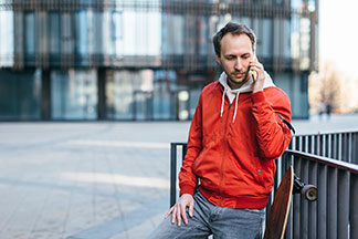 Športové bundy pre všedný deň: mladý muž v červenej športovej bunde s longboardom telefonuje na svojom smartfóne.
