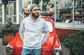 Mladý muž vo vlnenej čiapke a sivej mikine s kapucňou sa ležérne opiera o červené auto.