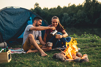 Správne vybavenie: mladý pár v outdoorovom oblečení sedí pri dobrej nálade pred svojím stanom pri ohni a pije kávu.