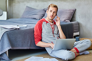 Mladý muž oblečený v súprave domáceho oblečenia sedí na podlahe opretý o posteľ, telefonuje a pracuje na svojom notebooku.